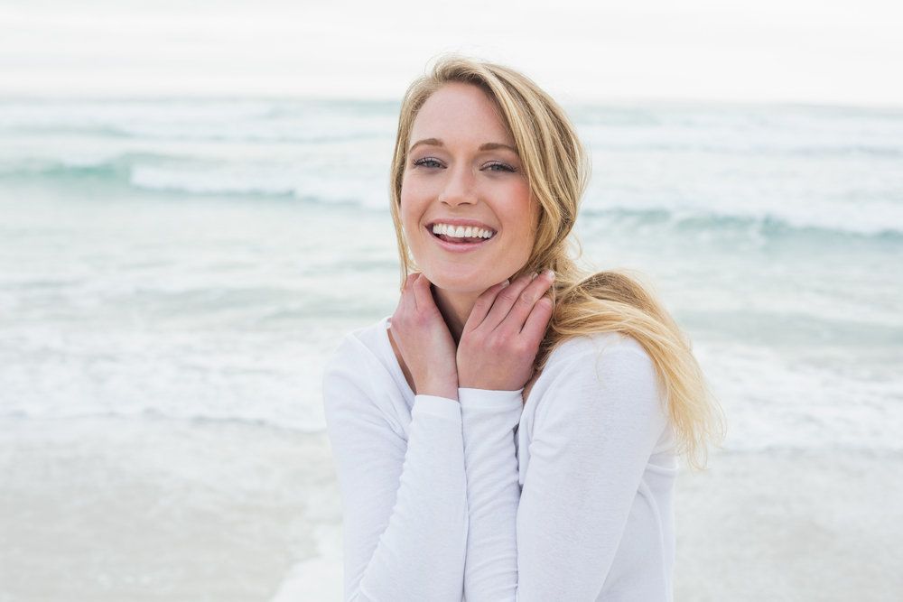 A beautiful woman with blonde hair wearing a white long-sleeved shirt smiles joyfully on the beach.